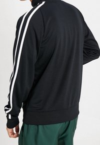 Hombre vestido con una chaqueta de chándal negra con dobles rayas blancas en las mangas, combinada con pantalones verdes, visto desde atrás y de lado.