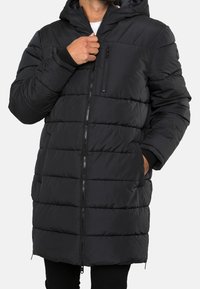 Schwarz gepolsterter Mantel mit einem durchgehenden Reißverschluss, Kapuze und elastischen Bündchen. Verfügt über eine Brusttasche und zwei Seitentaschen. Glattes Material mit einem gesteppten Design.