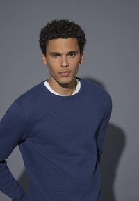 Blue textured sweater with a round neckline, worn over a white shirt. The model has short curly hair and a neutral background.