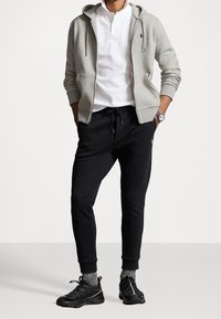 Felpa grigia con zip e tasche, polo bianca sotto, pantaloni jogger neri e sneakers nere con suole testurizzate e calzini a fantasia.