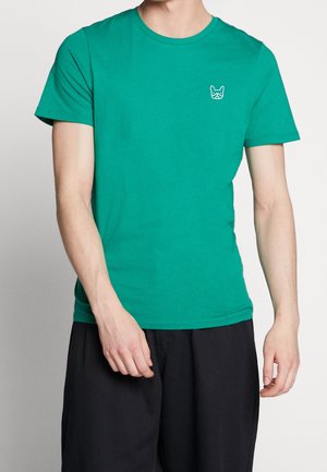 Homme portant un t-shirt vert à manches courtes avec un petit logo de chien blanc sur le côté gauche de la poitrine, assorti à un pantalon noir ample.