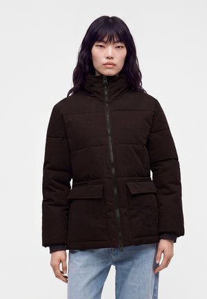 Junge Frau mit langen schwarzen Haaren trägt eine dunkelbraune gesteppte Jacke mit Reißverschluss und hellblaue Jeans vor einem schlichten Hintergrund.