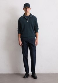 Dunkelgrüner Reißverschluss-Pullover, marineblaue Hose, schwarze Schuhe und passende Mütze. Der Pullover hat eine strukturierten Oberfläche, und die Hose ist tailliert mit flachem Bund.