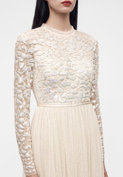 Femme portant une robe beige à manches longues avec des motifs complexes de sequins et perles blancs sur le corsage et les manches, et une jupe plissée.