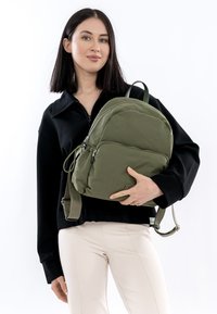 Groene nylon rugzak met een afgeronde vorm, dubbele rits en verstelbare bandjes, gedragen door een persoon die een zwart jack en lichte broek draagt.