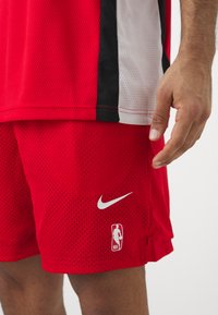 Røde netballshorts med en hvid Nike Swoosh og NBA-logo. Har åndbart stof og elastisk talje. Detaljeret syning.