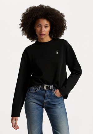 Schwarzes Langarm-T-Shirt aus Baumwolle, mit einem kleinen weißen gestickten Logo auf der linken Brust, kombiniert mit blauen Denim-Jeans und einem silbernen Gürtel mit Schnalle.
