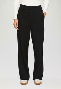 Pantalons noirs à jambes larges avec une texture lisse, deux poches latérales et une taille mi-haute, associés à des chaussures de couleur claire.