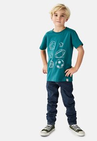 Tyrkis bomuldst-shirt med hvidt sportsinspireret motiv, kombineret med mørkeblå denimjeans og grønne høje sko med hvide snørebånd. Afslappet pasform.