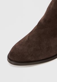 Botim de tornozelo em camurça castanha com bico apontado e textura suave. Apresenta uma sola de borracha preta minimalista, realçando um design elegante.