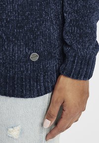 Mørkeblå teksturert genser med ribbestrikkede mansjetter og en sølvfarget logo-merke, kombinert med lyse denimbukser med et slitt detaljer på hoften.
