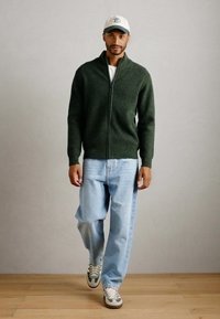 Mann med hvit caps, mørkegrønn zip-up genser, lyseblå jeans og hvite sneakers, gående på tregulv mot en ensfarget vegg.