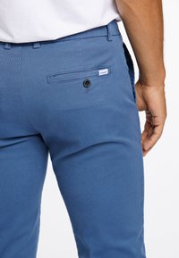 Blå bomullsbukser med en bakficka som har en svart knapp och en liten etikett. Tyget har en mjuk textur och en skräddarsydd passform.