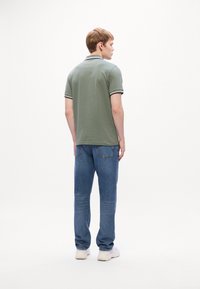 Ung mand står med ryggen til iført grøn polo, blå jeans og hvide sneakers mod en ensfarvet hvid baggrund.