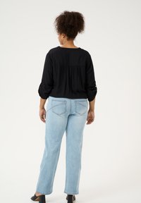Blusa negra de manga larga con detalle fruncido en la parte posterior, combinada con jeans rectos de color azul claro. El fondo es liso y de color claro.