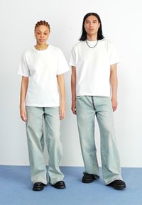Dos personas están de pie una al lado de la otra, vistiendo camisetas blancas de corte holgado y jeans azul claro desgastados, complementados con zapatos negros gruesos.
