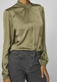 Olivgrön satinblus med smockad hals, långa ärmar och rynkade veck, i kombination med mörkgrå högmidjade jeans.