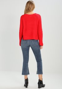 Pull rouge côtelé à manches longues, associé à un jean bleu évasé et des bottes à talons noires, vu de dos.