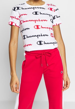 Person iført hvid kortærmet skjorte med gentagne røde og sorte "Champion" logoer og røde joggingbukser med hvide sidestriber og snøre.