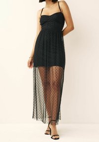 Schwarzes Pünktchenkleid mit taillierter Oberkante, transparenter �Überlagerung und verstellbaren dünnen Trägern. Akzentuiert mit einem fließenden Saum und schwarzen Sandalen.