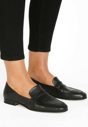 Mujer con mocasines negros de cuero sin cordones combinados con pantalones negros recortados, de pie sobre una superficie blanca.
