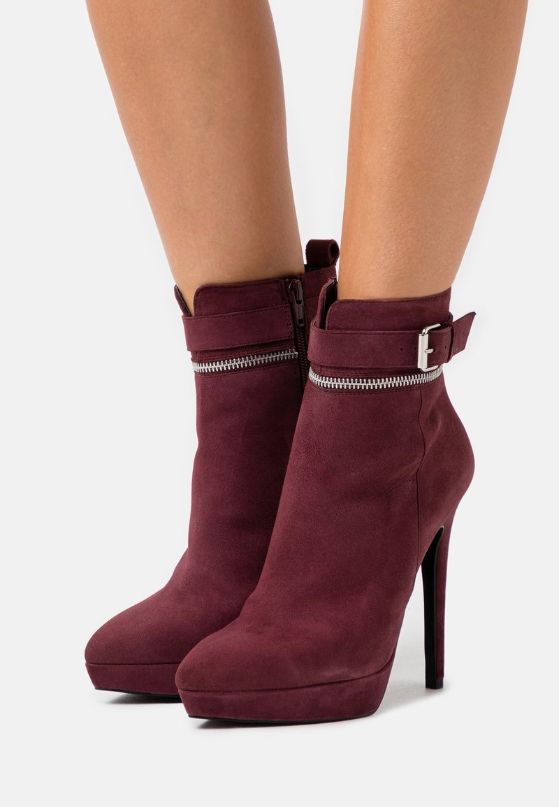 Bottines en daim rouge foncé pour femme avec hauts talons aiguilles, semelles plateformes, fermetures éclair latérales et brides à boucle autour des chevilles.