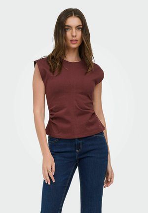 Kvinde med brunt hår iført en ærmeløs vinrød top og blå jeans, stående mod en ensfarvet lys baggrund.