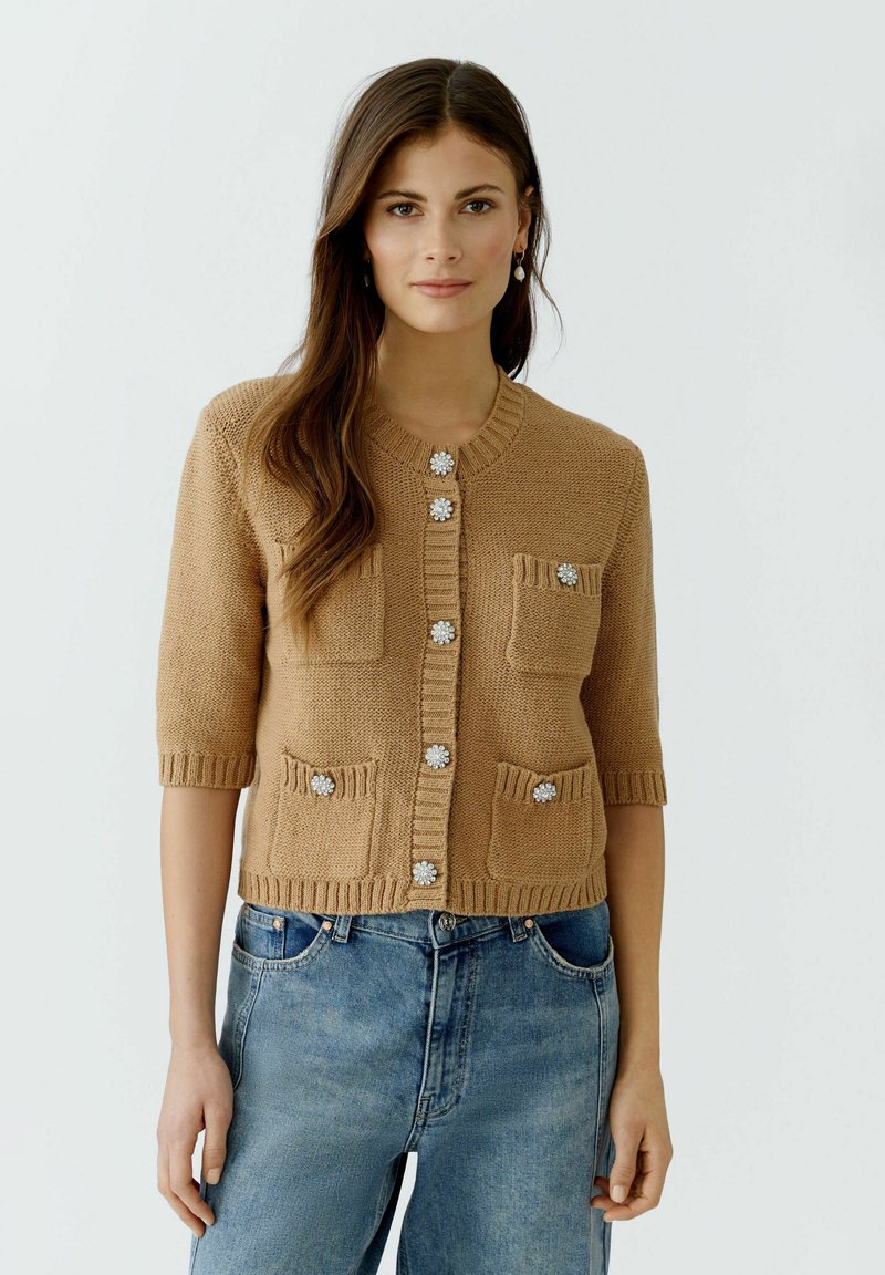 Brauner, gestrickter Cardigan mit kurzen Ärmeln, ausgestattet mit zwei Fronttaschen und dekorativen Blumenknöpfen, getragen über blauen Jeans.