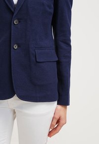 Blazer bleu marine en tissu texturé, fermeture à un bouton, et poche poitrine gauche, porté par-dessus une chemise blanche et un pantalon blanc.