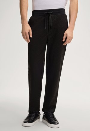 Homme portant un pantalon noir texturé à cordon, des baskets noires à semelles blanches et une chemise blanche rentrée, debout devant un fond uni.