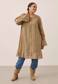 Femme portant une robe ample beige par-dessus un jean bleu et des bottes de cow-boy beige, debout, une main touchant son cou et souriante.