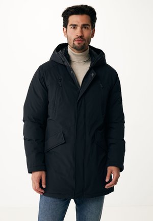 Zwarte pufferjas met een capuchon, voorzien van twee borstzakken, zijzakken en een ritssluiting. Gestaan over een lichtbeige coltrui.