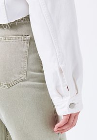 Valkoinen farkkutakin hiha nappi-yksityiskohdalla, yhdistettynä vaaleanvihreisiin samettihousuihin, joissa on takatasku ja pehmeä ribbikuvio.