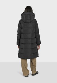 Zwarte donzen jas met een capuchon, met een gewatteerde structuur, een getailleerde taille en een lange pasvorm. Draagt over beige broeken en stevige zwarte schoenen.