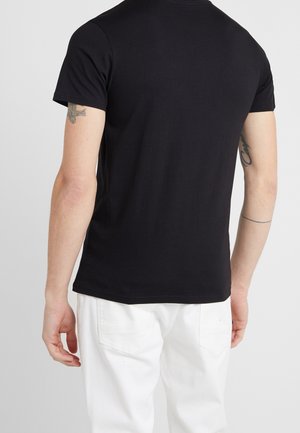 Schwarzes kurzärmliges T-Shirt aus Baumwolle mit rundem Ausschnitt und schlichtem Rückendesign. Weiße Hose darunter gezeigt.