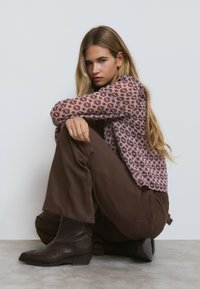 Suéter de punto con un patrón geométrico morado, pantalones anchos marrones y botas de estilo western marrones con tacón bajo, sentado contra una pared blanca.