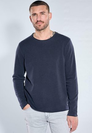 Man with short hair and beard wearing navy long-sleeve shirt and light gray pants, standing against a plain light background.