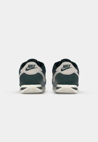 Nike tenisice s gornjim dijelom od tamnozelene nubuk kože, s sivim detaljima, bijelom srednjom sole i crnom oznakom na peti, prikazane sa stražnje strane.