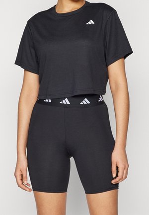 Kobieta ubrana w czarny krótki t-shirt i dopasowane wysokie szorty rowerowe z białymi logo Adidas na pasie i na piersi.