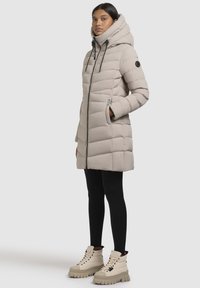 Manteau doudoune beige avec un col haut et une fermeture éclair sur le devant, présentant une texture matelassée et des accents noirs, associé à des leggings noirs et des bottes crème.