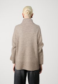 Beige Strickpullover mit hohem Rippkragen, lockerer Passform und gerippten Bündchen. Glatte Textur mit leichtem Schimmer und subtiler Farbschattierung.