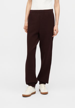 Personne portant un pantalon de jogging marron foncé coupe décontractée avec des poignets élastiques et des baskets blanches, debout devant un fond uni.