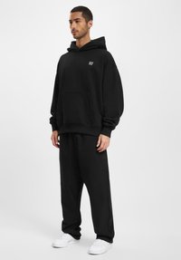 Czarny, oversize hoodie z kieszenią typu kangur i białym napisem "DEF" na klatce piersiowej, w połączeniu z luźnymi czarnymi spodniami dresowymi i białymi sneakersami.