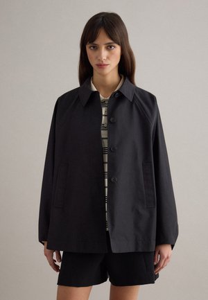 Donna che indossa una giacca nera con bottoni sopra una camicia a righe e pantaloncini neri, in piedi davanti a uno sfondo neutro uniforme.