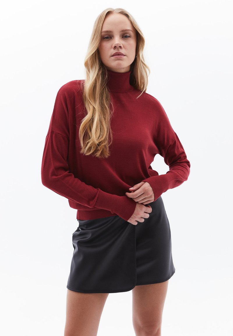 Maglione rosso a maniche lunghe con collo alto, caratterizzato da una vestibilità rilassata e polsini a coste, abbinato a una mini gonna nera in finta pelle.