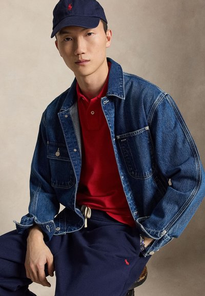 Νέος άνδρας που φοράει καπέλο μπλε ναυτικού, κόκκινο πόλο μπλουζάκι, μπλε τζιν μπουφάν και μπλε σκούρα φούτερ παντελόνια, καθισμένος μπροστά σε απλό μπεζ φόντο.