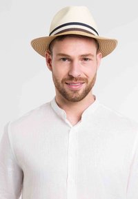 Sombrero de paja de color beige claro, con un ala ligeramente levantada y una banda a rayas en negro y marrón. Combinado con una camisa blanca de lino con botones.