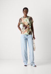 Blomstermønstret t-skjorte med brun bakgrunn, parret med lyseblå vidde jeans og matchende åpne hæler, holder en kremfarget håndveske.
