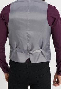Gilet grigio satinado con dettaglio della fibbia sul retro, abbinato a una camicia a maniche lunghe di un profondo viola e pantaloni neri. Tessuto liscio, vestibilità su misura.