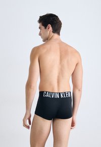 Schwarze Boxershorts mit einem breiten weißen "CALVIN KLEIN" Logo-Bund, die eng anliegen und eine glatte Textur aufweisen. Modell von hinten gezeigt.